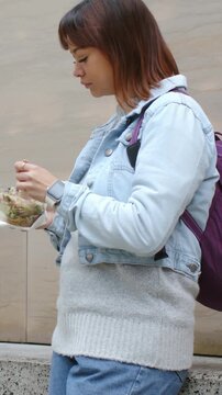 Vertical video: Holding takeaway box and stirring, woman eating by bench with fork for lunch