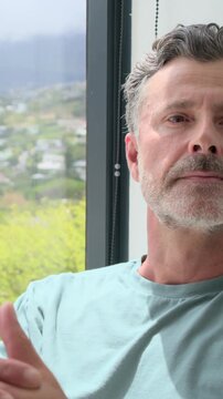 Vertical video: Camera moving closer, man turning head drifting right by window at home in teal tee