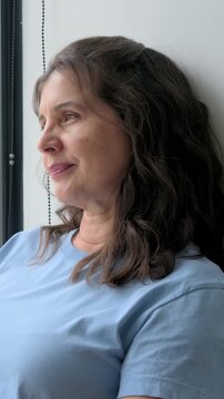 Vertical video: Sitting lady in blue tee looking, turning, laughing by window with light shifting