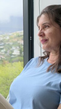 Vertical video: Shifting as camera zooming woman blue-top beige-pants smiling at window, copy space