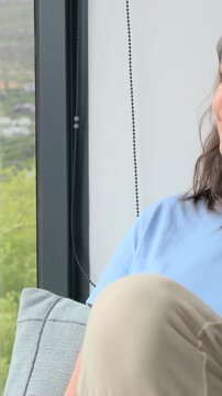 Vertical video: Glancing at window, woman in blue tee relaxing drawing knee on sofa, copy space