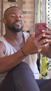 Vertical video: Adjusting man in tank angling maroon phone for selfie in gym brick wall bracelet