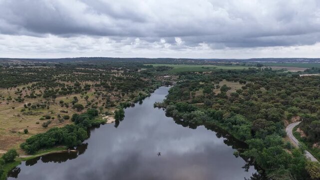 Aerial view of a winding river flowing through lush green hills with a small dam and ecological barrier under a cloudy sky in Portugal.