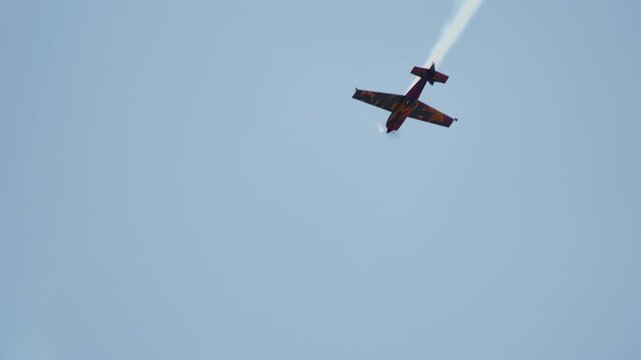 Vivid red and black patterned aerobatic monoplane in a steep banked turn with smoke trail visible against light blue sky viewed from below
