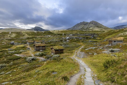 Wooden huts in an open meadow in a remote mountain landscape, with winding paths and clouds in the sky, Peer Gynt hytta, Rondane National Park, Otta, Innlandet, Norway