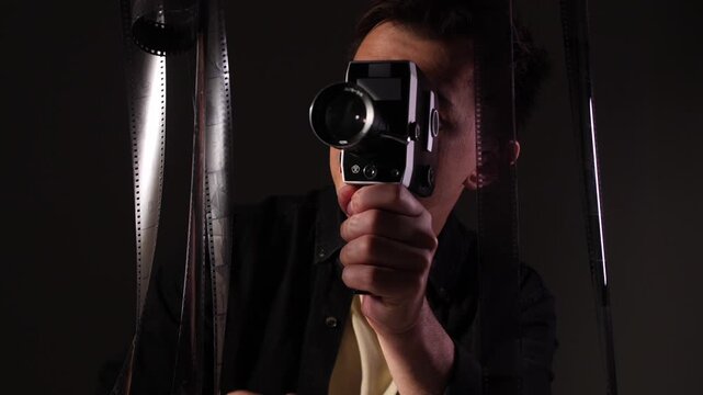 Filmmaker holding an old movie camera, actively shooting a scene, surrounded by hanging celluloid film strips representing the creative process of traditional cinema and analog film production