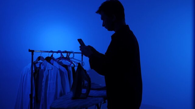 Man in silhouette standing by an ironing board and clothes rack, distracted by his smartphone instead of ironing laundry, highlighting digital distraction during household chores