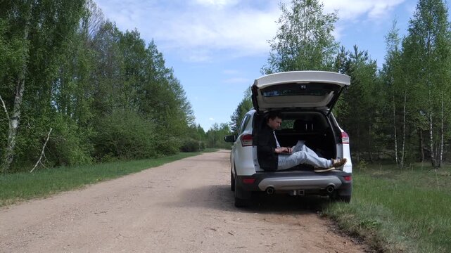 Man sitting in the open trunk of an suv, actively working on a laptop while parked on a dirt road surrounded by green trees, embracing a flexible digital nomad lifestyle