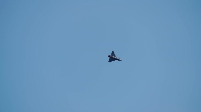 Tiny dark silhouette of a modern delta wing canard military fighter jet crosses a vast clear blue sky during an aerial display event