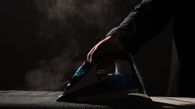 Hand holding an iron pressing a garment on an ironing board, creating a cloud of steam, highlighting daily household chores and laundry tasks against a dark background