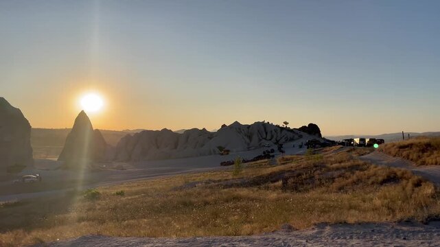 Stunning 4K wide-angle shot of the sun setting behind iconic fairy chimney rock formations in Cappadocia, Turkey. Warm golden light bathes the volcanic tuff landscape as lens flare radiates