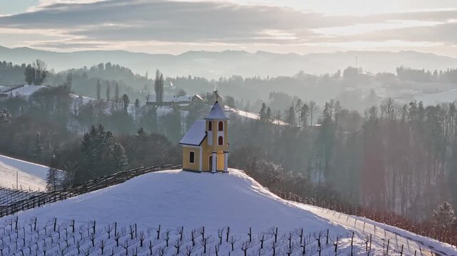 Aerial view of Dreisiebner Kapelle standing on a snow-covered vineyard hill surrounded by a misty winter landscape under soft light in Gamlitz, Styria, Austria.