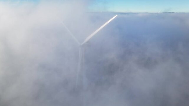 Aerial view of Windpark Freilanderalm with wind turbine blades emerging from thick white clouds under a clear blue sky in Modriach, Styria, Austria.