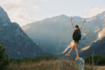 Young woman with backpack stands on a rocky ledge, arms raised in celebration, surrounded by rugged mountains and a clear blue sky, her casual outfit and excited expression conveying freedom and © SHOTPRIME STUDIO