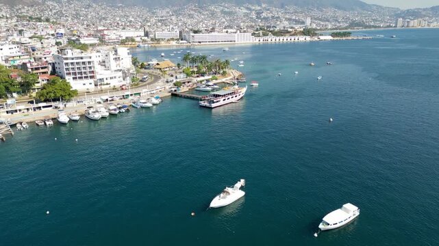 Captivating aerial drone footage showcasing Acapulco Bay in Mexico, with boats dotting the tranquil water and the vibrant coastal city extending to the lush green hills under clear skies. Ideal for tr