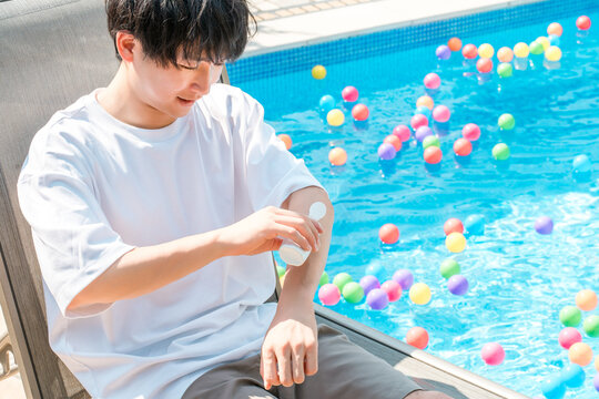 A young Asian man applies sunscreen to his arm by the poolside.