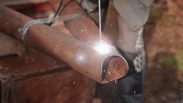 Detailed view of electric welding on metal pipe in backyard workshop setting. Skilled homeowner uses protective gloves and modern equipment for durable steel construction repair outdoors.