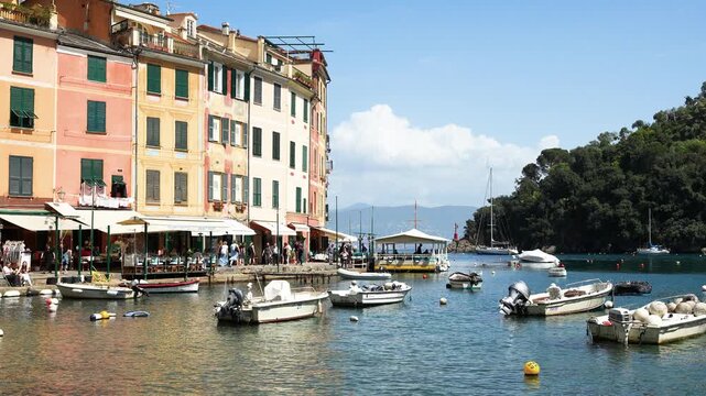 Portofino harbor sparkles under bright sunlight, charming pastel buildings line the waterfront, small boats bob in tranquil blue water, lush green hillsides behind, vibrant and inviting scene.