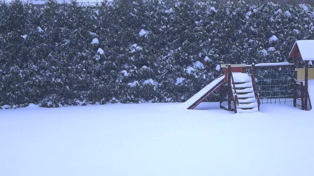 Quiet, deserted children's playground in a backyard getting completely covered by heavy snowfall during a cold and desolate winter day, creating a peaceful and serene seasonal scene