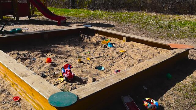 Brightly colored plastic toys left scattered in a large wooden sandbox at an empty outdoor playground on a sunny day, with sand being gently blown by the wind, evoking a feeling of abandonment