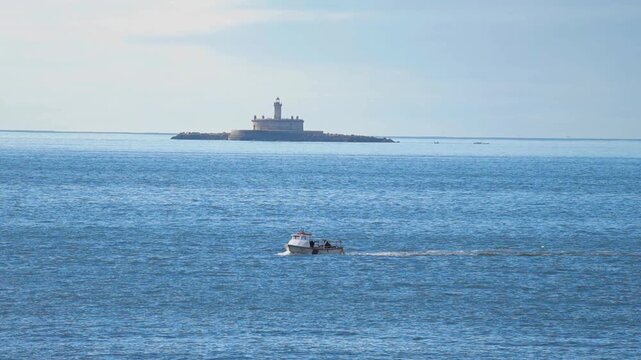 Small fishing boat sailing across the tranquil blue waters of the Tagus river estuary with the historic Bugio Lighthouse visible on a distant island under a hazy sky in Portugal