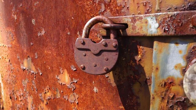 Close up view of a weathered brown padlock on a rusted hasp, securing an old metal door with peeling paint and significant corrosion damage, moving in a slow pan on a sunny day