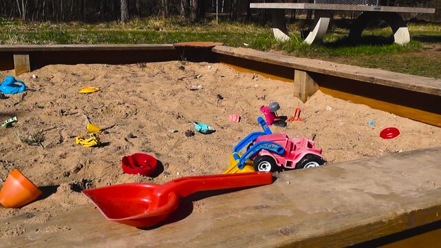 Colorful plastic toys like a shovel and a dump truck are scattered in an empty wooden sandbox at an outdoor playground on a sunny day, suggesting the end of playtime or a forgotten play area