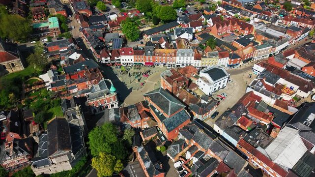 Top down aerial drone footage of Romsey town centre in Hampshire, showing historic streets, rooftops, shops, roads, public spaces, pedestrians, and surrounding town buildings on a sunny day.