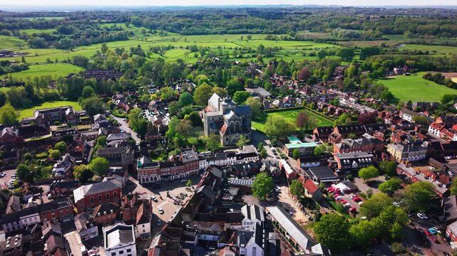 High aerial drone footage of Romsey Abbey in Hampshire, showing the historic abbey church, town centre, rooftops, streets, green spaces, trees, and surrounding countryside on a sunny day.
