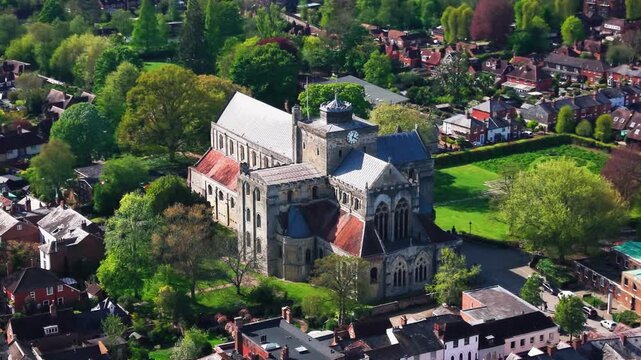 Aerial drone footage of Romsey Abbey in Hampshire, showing the historic abbey church, stone architecture, clock tower, surrounding trees, green spaces, rooftops, and town landscape on a sunny day.
