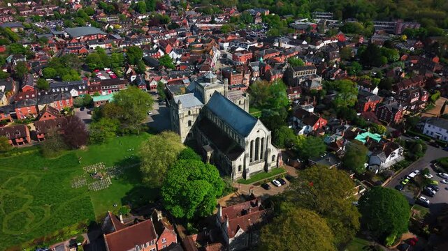 High aerial drone footage of Romsey Abbey in Hampshire, showing the historic abbey church, churchyard, surrounding rooftops, streets, trees, green spaces, and wider town centre on a sunny day.