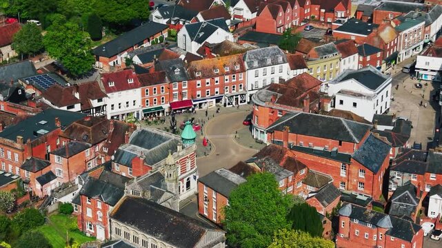 Aerial drone footage of Romsey town centre in Hampshire, showing the high street, historic buildings, rooftops, shops, roads, pedestrians, and local architecture on a sunny day. Useful for British