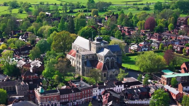 Wide aerial drone footage of Romsey Abbey in Hampshire, showing the historic abbey church, surrounding town centre, rooftops, green spaces, trees, streets, and countryside beyond on a sunny day.