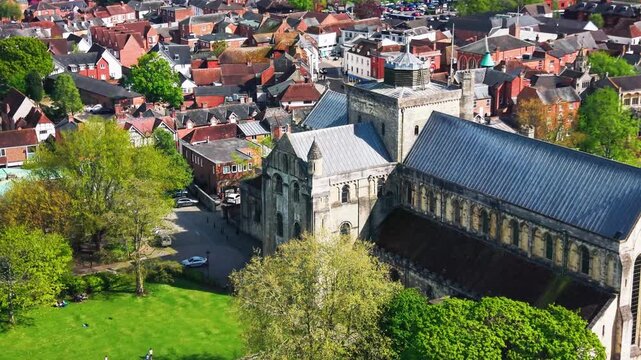 Close aerial drone footage of Romsey Abbey in Hampshire, showing the historic abbey church, stone architecture, roof detail, surrounding streets, town buildings, trees, and green spaces on a sunny day