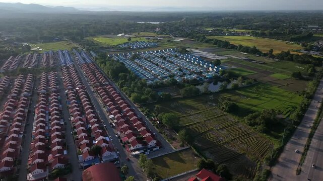 An expansive aerial view showing the intersection of dense, uniform red-roofed housing blocks and agricultural rice fields. The shot highlights the encroaching development in a scenic valley