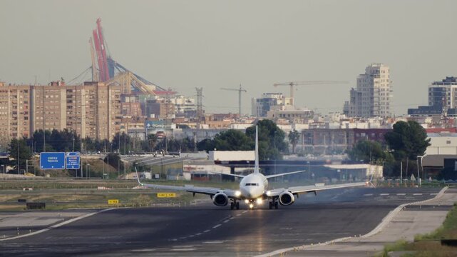 White narrow body commercial jet rolls on runway facing camera with landing lights on, city skyline and port cranes visible through morning haze in background