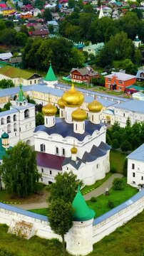 Vertical video. Russia, Kostroma. Holy Trinity Ipatievsky Monastery, Drone footage over white stone walls, golden domes and green rooftops along the riverbank in summer. Drone footage, Point of inter