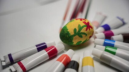 A vibrantly decorated Easter egg rests in the center of a scattered array of colorful paint tubes and art brushes on a white background. © Ahmad Khan