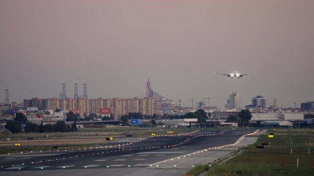 White commercial aircraft descends on final approach over an illuminated runway at dusk, city skyline with port cranes and pink-purple sky in background