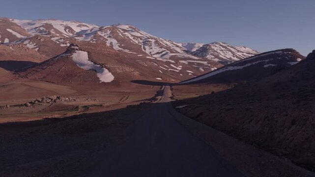 Straight empty asphalt road cutting through a vast arid Atlas Mountains valley with snow patches