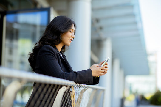 Woman looks at smartphone while standing by railing outside office building in daylight