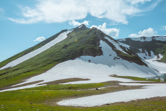 Scenic alpine view to sunlit big hill with green hillside, glacier and sharp rocky peak under clouds in blue sky. White tent on small islet among glacial lake and snows in high mountains in sunlight.