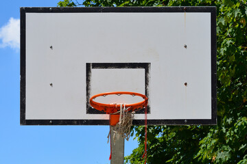An outdoor basketball backboard with an orange hoop and a damaged torn net stands against a clear blue sky and green trees © nahhan
