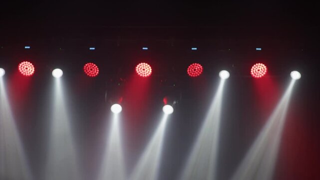 Concert lighting rig viewed from below at diagonal angle, white spotlight beam and multiple red LED par fixtures visible, blurred crowd and warm stage glow at left