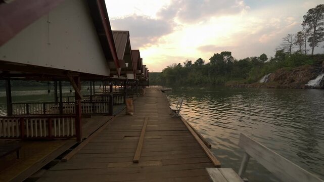 POV walking shot along a wooden floating dock beside Tad Khoo waterfall, with riverside huts, benches and golden hour light on the water.