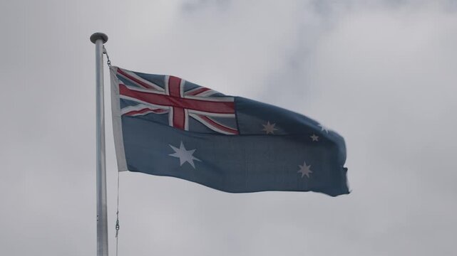 An Australian flag blowing in the wind on top of a high flag pole with cloudy grey sky in the background.
