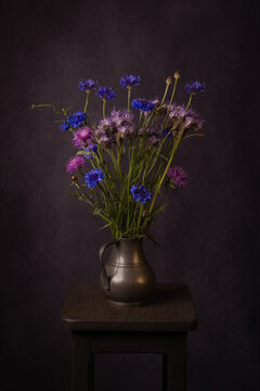 stilllife in painterly renaissance style with cornflowers in metal vase on stool