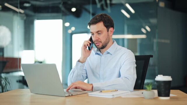 Serious male designer speaking on mobile phone and working on laptop computer. Side view of digital nomad sitting at corporate workspace desk with notebook, documents and disposable coffee cup.