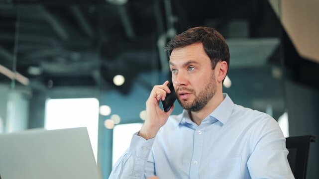 Focused male creator or digital nomad speaking on mobile phone and looking at computer monitor. Close-up portrait of professional specialist consulting client or working remotely at coworking desk.