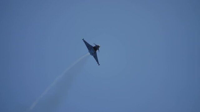 Military fighter jet performs a high speed aerobatic maneuver against a clear blue sky. The aircraft climbs, banks, and accelerates with afterburners glowing, leaving a vapor trail
